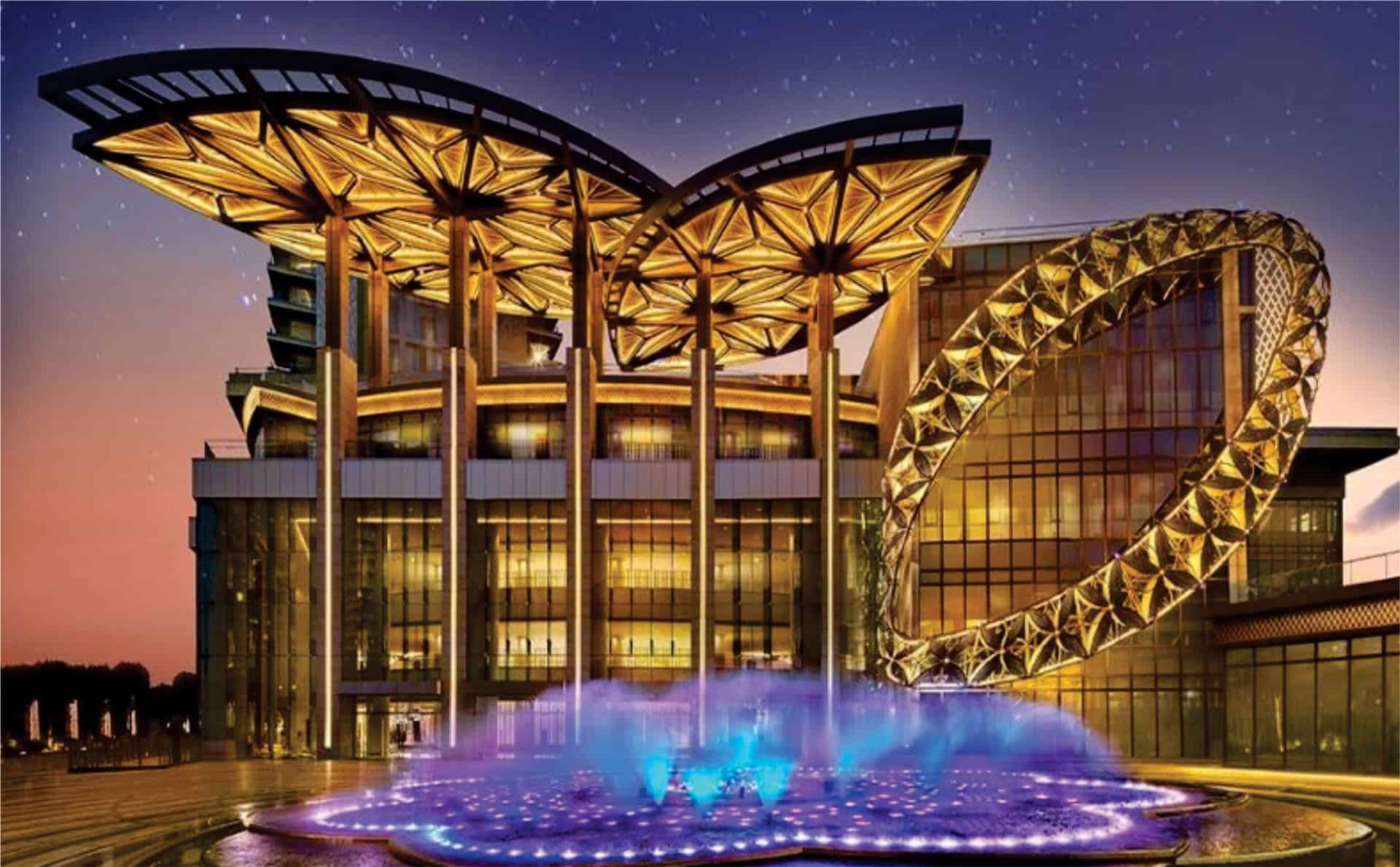 Luxury hotel with illuminated architecture and fountain at sunset, featuring modern design and vibrant lights.