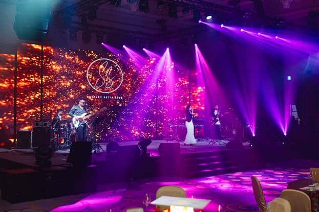 Live band performing with colorful stage lights at an event, featuring a vibrant digital backdrop display.